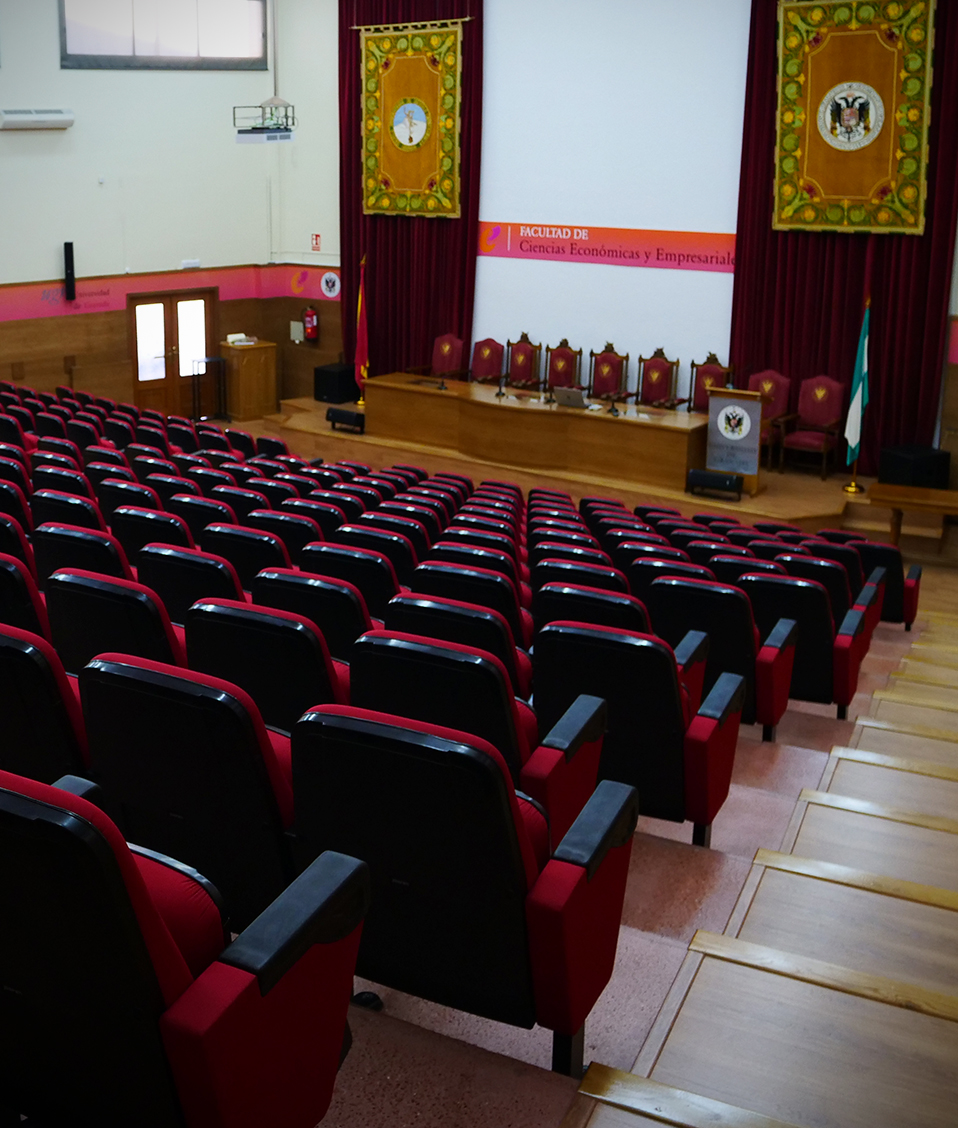Sala para dar discursos y hacer presentaciones en la Facultad de Ciencias Económicas y Empresariales de Granada. Al fondo se ven cortinas con el logo de la Universidad de Granada y en primer plano hay butacas de color rojo